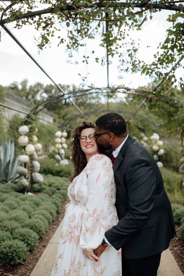 Tristan kissing Hailey on the cheek as she laughs, beneath the garden arbor