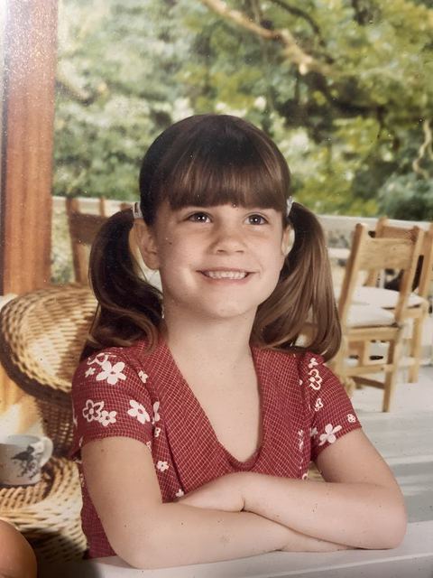 Hailey as a girl in a red top, arms crossed and smiling