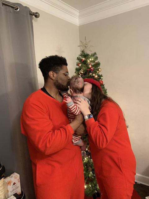 Tristan kissing Hailey while she holds their newborn by the Christmas tree