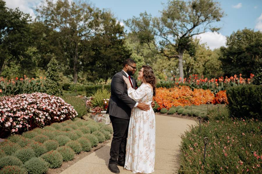 Hailey and Tristan embracing on a garden path surrounded by beds of autumn flowers